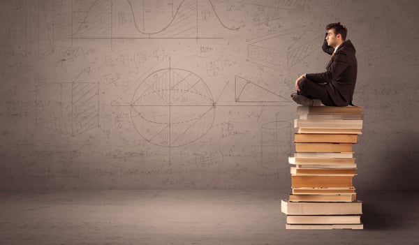 A serious businessman in suit sitting on a pile of giant books in front of a greyish brown wall with drawn lines, angles, numbers, circles and curves.